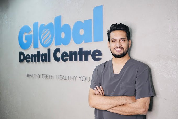 Dr. Nimesh Jain standing with arms crossed in front of the Global Dental Centre wall logo, smiling confidently while wearing a gray dental scrub.