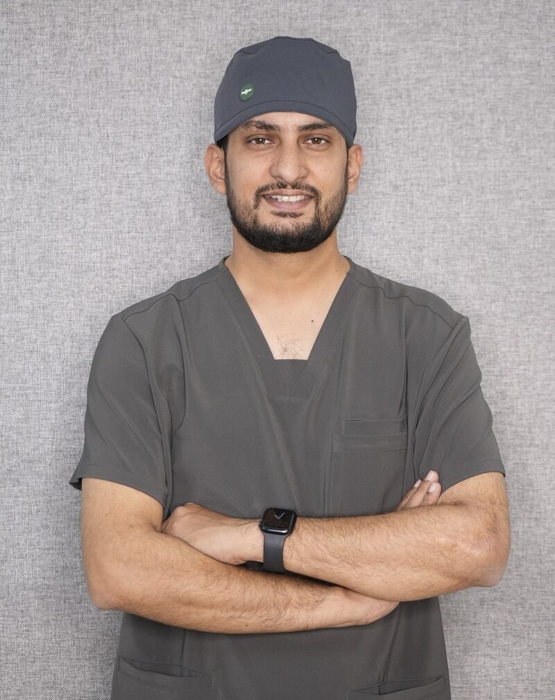 Dr. Nimesh Jain, a dentist and implantologist, standing with arms crossed, wearing a dark grey dental scrub and surgical cap, offering a friendly smile.