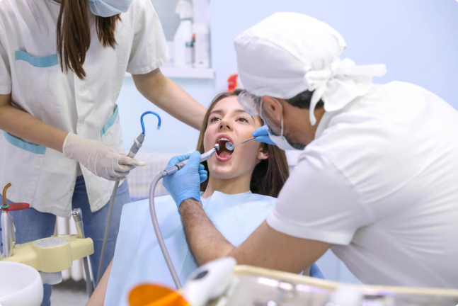 A dental team is performing a treatment on a patient in a dental clinic. One dentist is working on the patient's mouth while the assistant prepares tools.