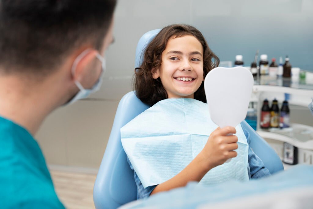 A young girl smiles and holds a dental mirror while sitting in a dentist’s chair during a dental visit.