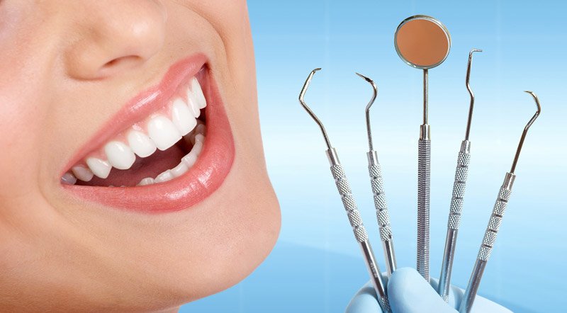 Close-up of a smiling person with bright white teeth, alongside dental tools such as a mirror, scaler, and probe, held by a gloved hand.