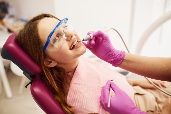 A young girl with protective glasses sits in a dental chair while a dentist with pink gloves uses a dental tool to clean her teeth.