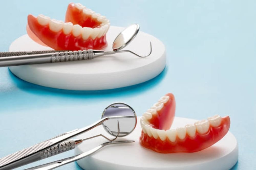 Two sets of dentures placed on circular white bases with dental tools, including a dental mirror and probe, positioned beside them on a light blue background.